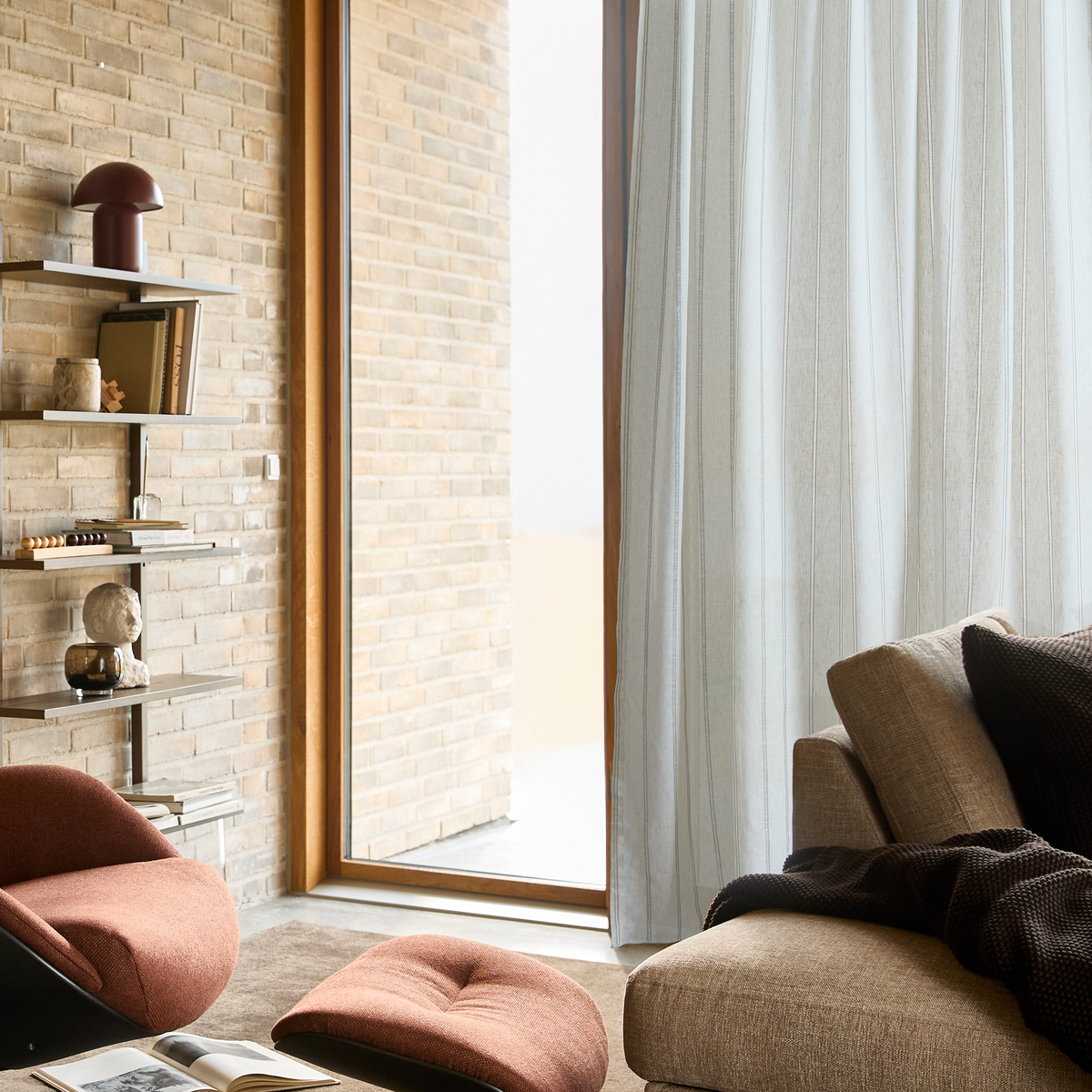 Lyst hjørne af stue med beige sofa med mørkt plaid og puder, let gardin ved stort vindue, væghylder med bøger, lille bordlampe og skulptur samt rustfarvet fodskammel på tæppe.