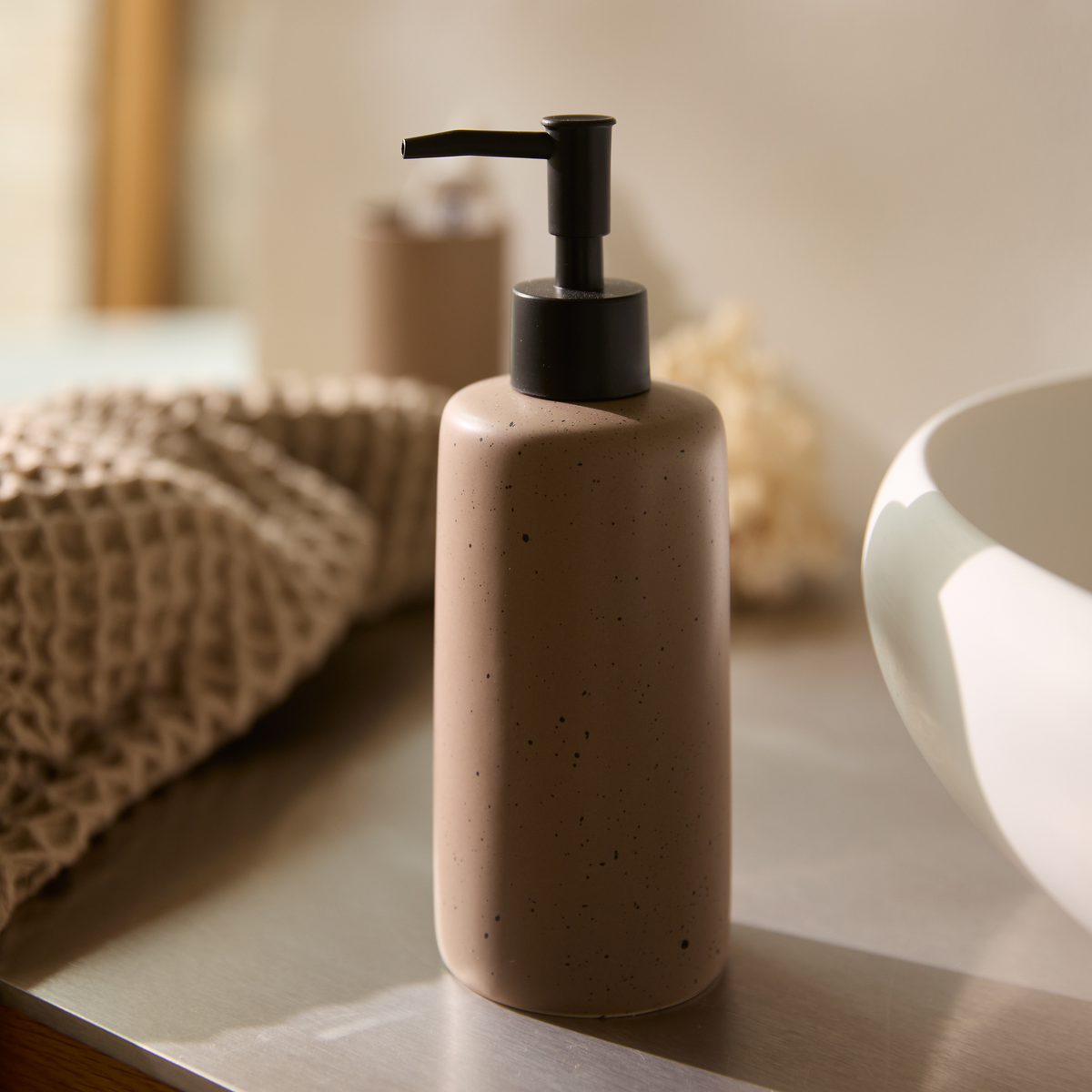 Beige sæbedispenser i keramik med sort pumpe på badeværelsesbord i blødt dagslys.