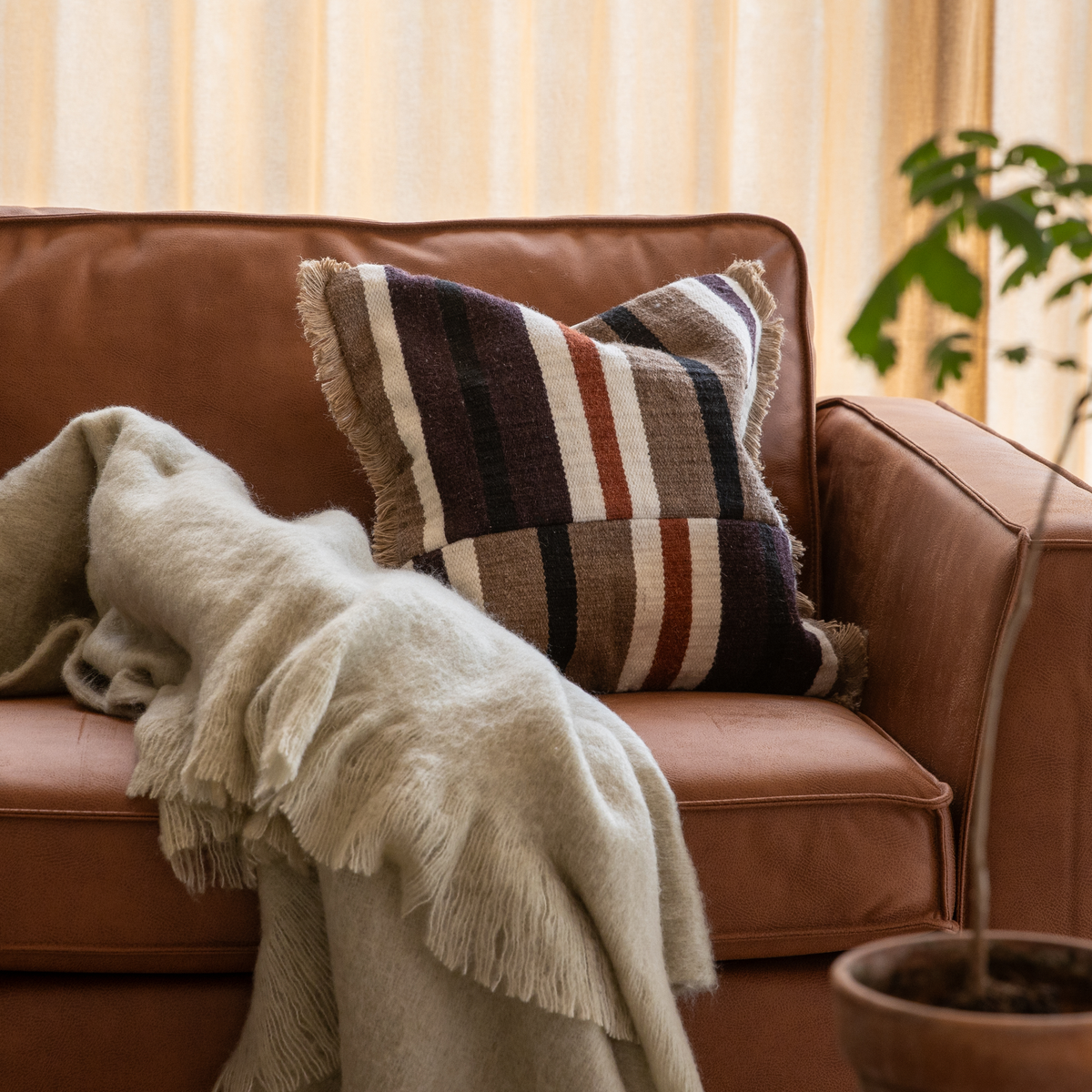 Brun lædersofa med stribet pude med frynser og en blød beige plaid; plante og lyse gardiner i baggrunden.