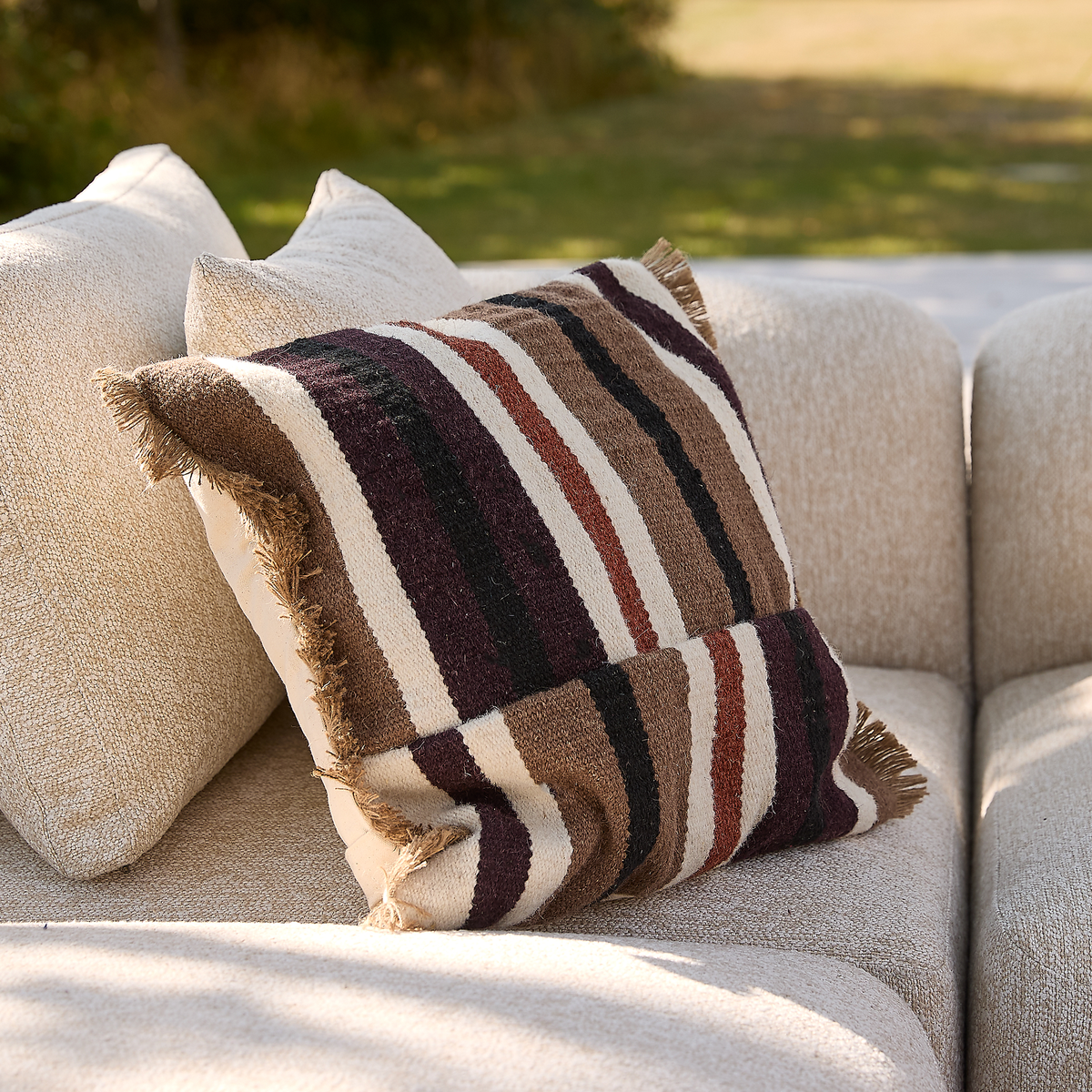 Stribet pude med frynser i brune, sorte, beige og vinrøde nuancer på en lys beige sofa udendørs i dagslys.
