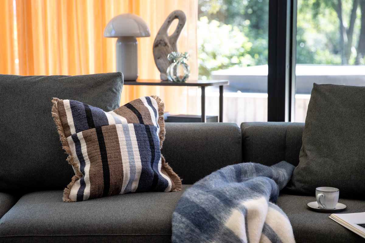 Mørkegrå sofa med stribet pude i blå, brun og hvid samt blåhvid plaid; lille espressokop på sædet, dagslys fra store vinduer og orange gardiner.