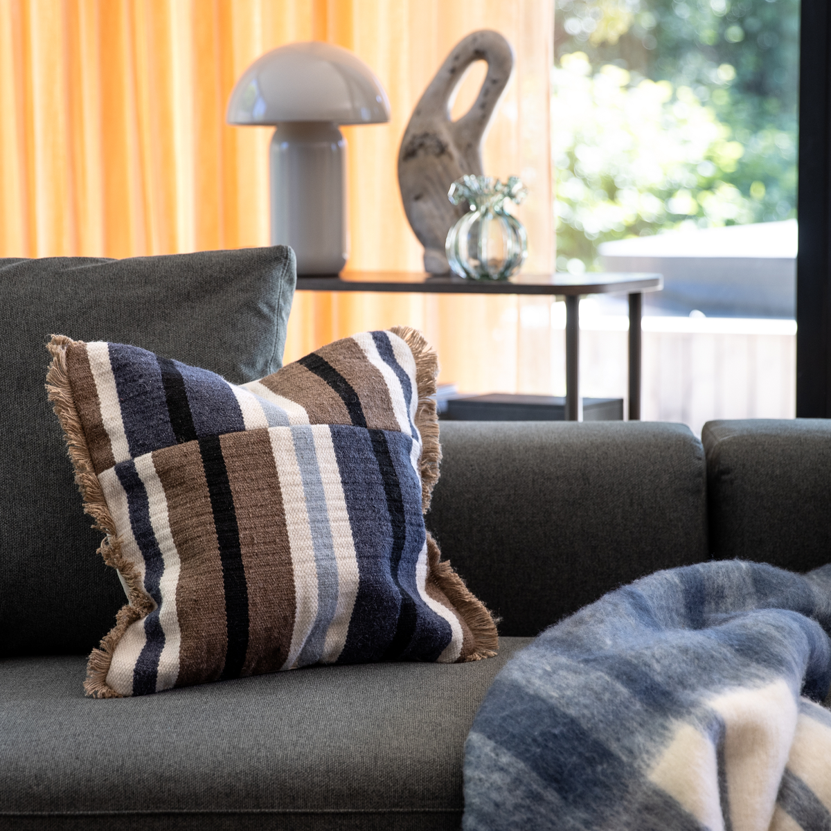 Stribet pude i blå, brun og hvid på en grå sofa med matchende plaid; i baggrunden et sidebord med hvid bordlampe, glasvase og skulptur foran varme gardiner i dagslys.