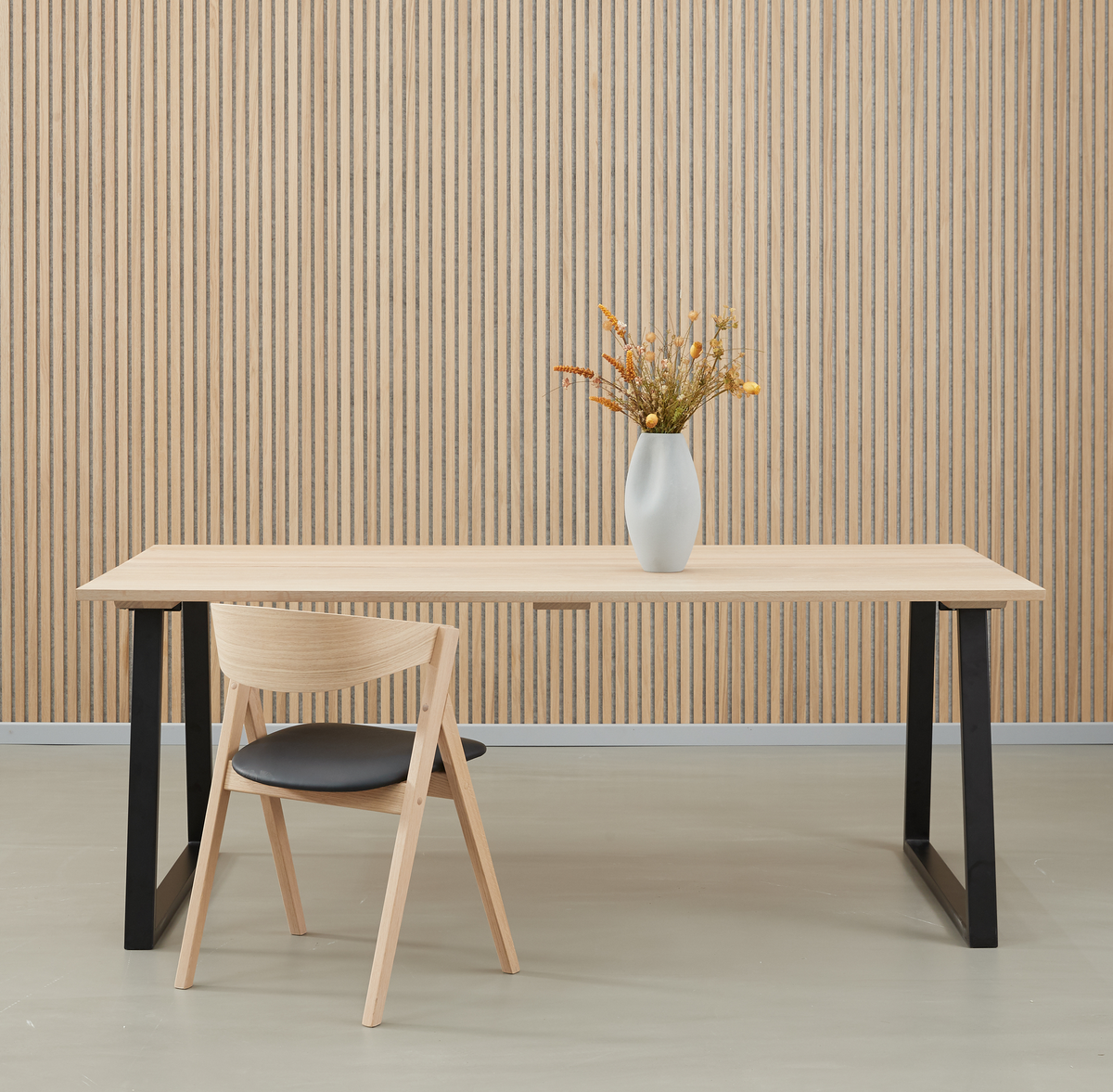 Minimalistisk rum med lyst træbord med sorte ben, en træstol og en hvid vase med tørrede blomster foran en væg med vertikale trælameller.