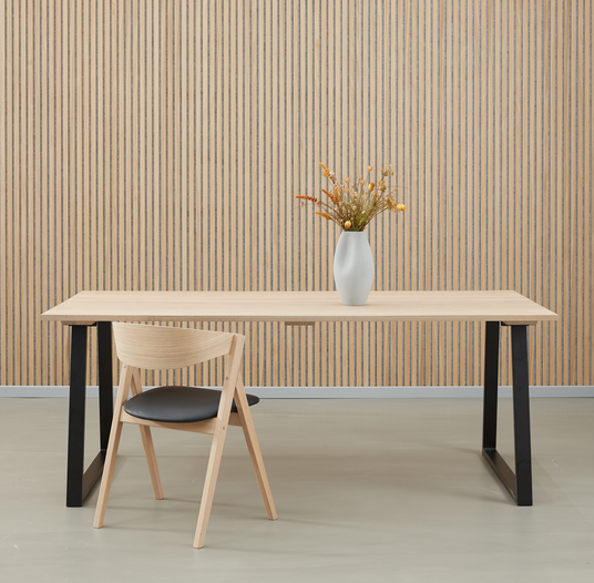 Minimalistisk rum med lyst træbord med sorte ben, en træstol og en hvid vase med tørrede blomster foran en væg med vertikale trælameller.