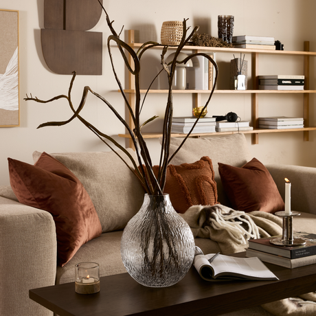 Beige stue med sofa, rustfarvede puder og plaid, glasvase med tørre grene på sofabord med tændt lys, bøger (bl.a. ”CHANEL”) og åben notesbog; træhylde og vægkunst i baggrunden.