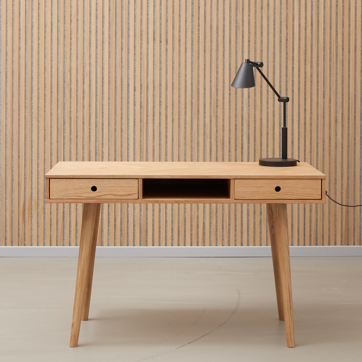 Minimalistisk skrivebord i lyst egetræ med to skuffer og et åbent midterrum, sort bordlampe ovenpå foran en lamelbeklædt væg.