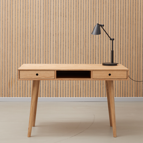 Minimalistisk skrivebord i lyst egetræ med to skuffer og et åbent midterrum, sort bordlampe ovenpå foran en lamelbeklædt væg.