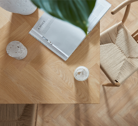 Top-down visning af hjørnet af et lyst egetræsbord med stol med flettet sæde, et opslået magasin med titlen "A SPACE TO GATHER", et terrazzo-objekt og et lille glas i blødt dagslys.