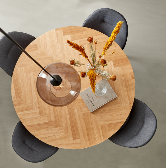 Rundt spisebord i lys eg med sildebensmønster, tre mørkegrå stole, glaspendel over, glasvase med tørrede blomster og bogen The Kinfolk Home på bordet.
