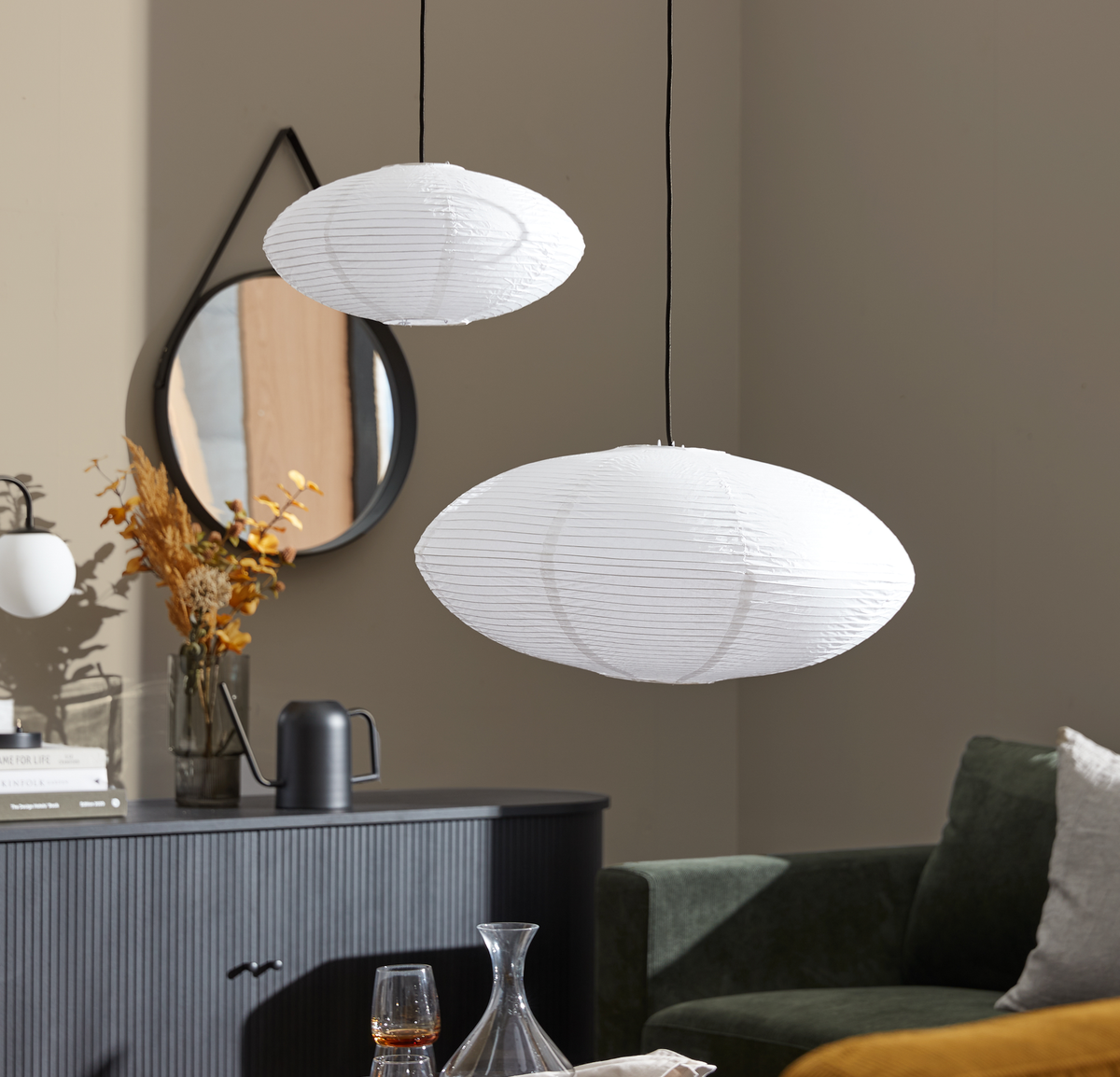 To hvide rispapir-pendeloplamper hænger over en stue med mørk skænk, rundt spejl og grøn sofa; en vase med tørrede grene og en bog med titlen ”For Life” står på skænken.