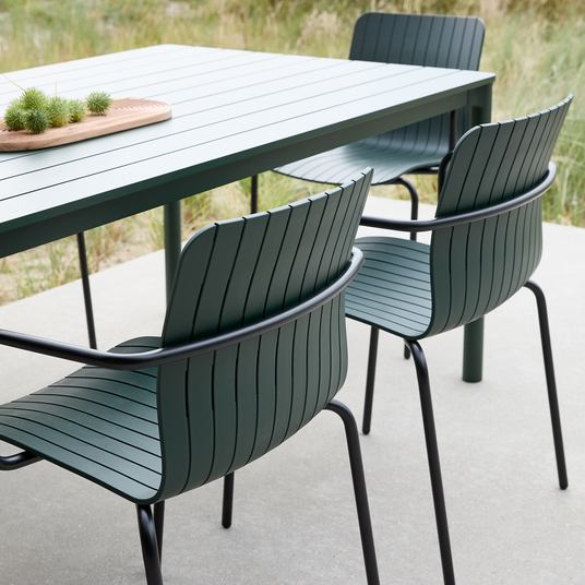 Grønt udendørs spisebord med lameller og matchende ribbede metalstole på terrasse; træbakke med små grønne planter på bordet.
