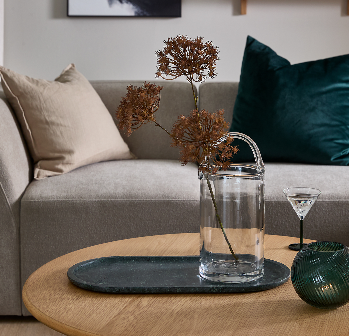 Beige sofa med puder bag et rundt sofabord i træ med mørk stenbakke, glasvase med tørrede grene og et lille cocktailglas.