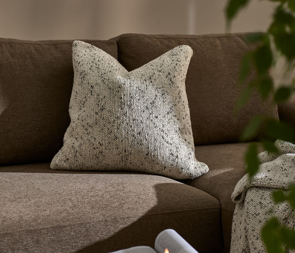 Strikket, spættet beige pude på brun sofa med matchende plaid i blødt dagslys.