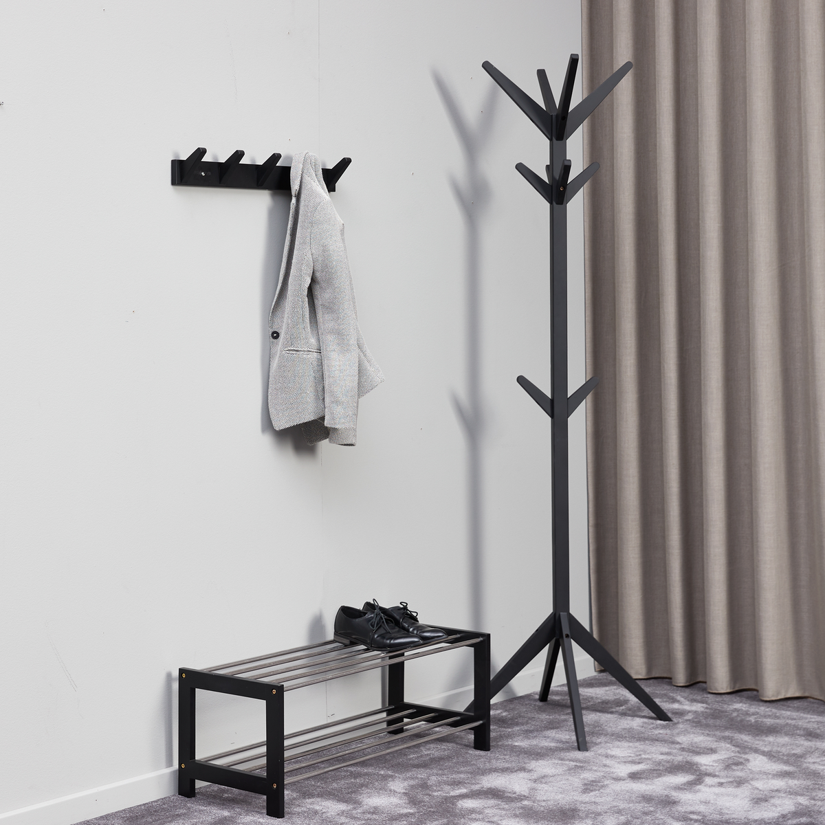 Minimalistisk entré med sort gulvstativ til tøj, vægmonteret knagerække med grå jakke og sort skoreol med et par sorte sko, gråt tæppe og beige gardin.