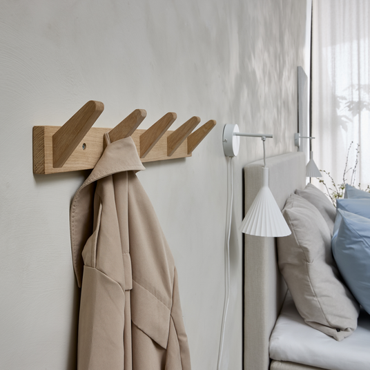 Vægmonteret knagerække i træ med beige frakke i et roligt soveværelse med hvid væglampe, neutrale puder og blødt dagslys.