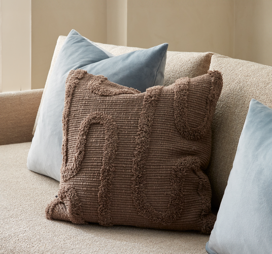 Nærbillede af brun, tekstureret pyntepude med tuftet mønster på en beige sofa, flankeret af to lyseblå velourpuder.