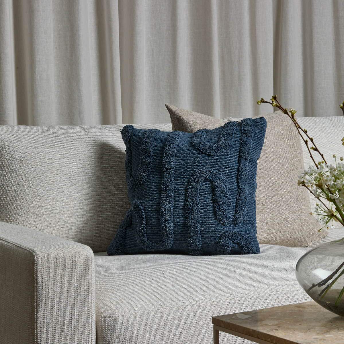Beige sofa med blå tekstureret pyntepude og beige pude foran lyse gardiner, vase med hvide grene på sofabord.