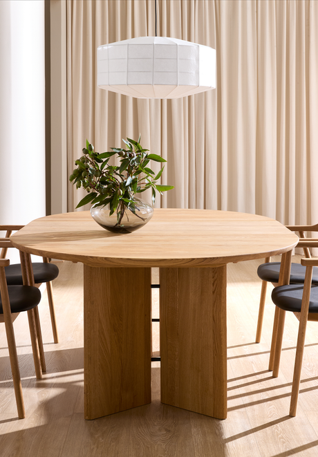 Rundt spisebord i eg med søjleunderstel omgivet af træstole med sorte sæder; en glasvase med grønne grene står på bordet og en hvid pendel hænger foran beige gardiner.