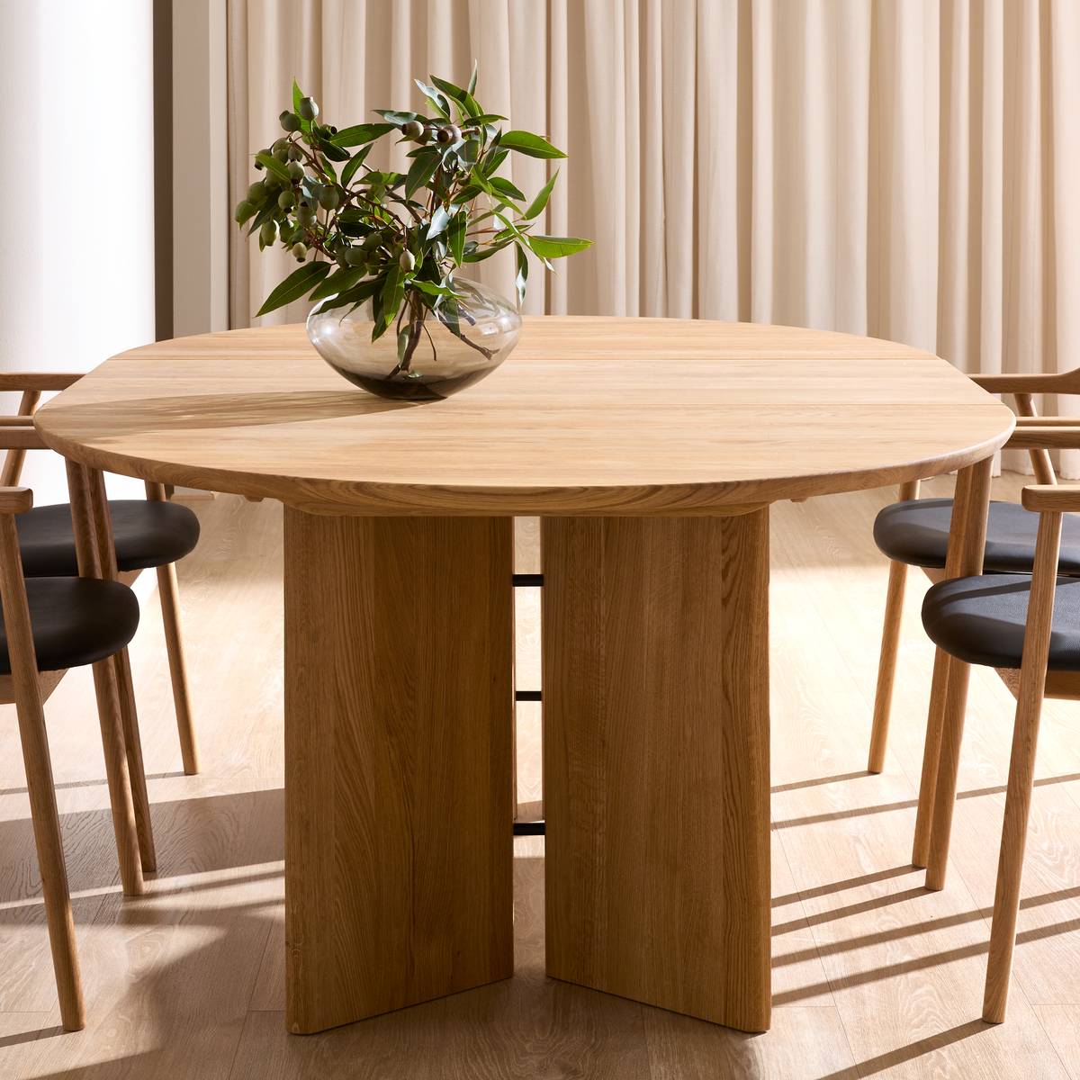 Rundt spisebord i lys eg med massiv sokkel, træstole med sorte sæder og en glasvase med grønne grene foran beige gardiner.