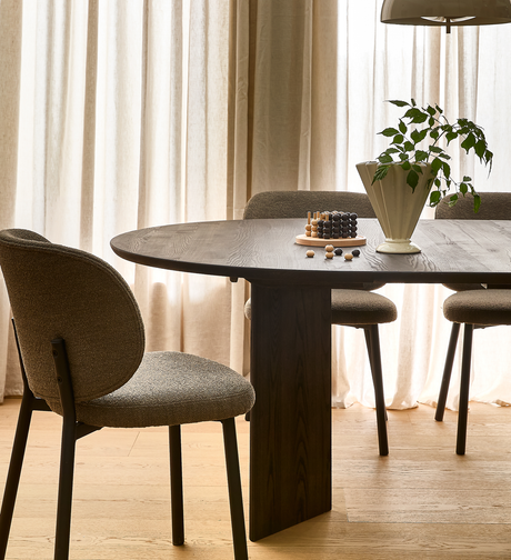 Mørkt, ovalt spisebord i træ med tre polstrede stole, beige gardiner i dagslys, en cremefarvet vase med grøn plante og et træspil med kugler på bordet.