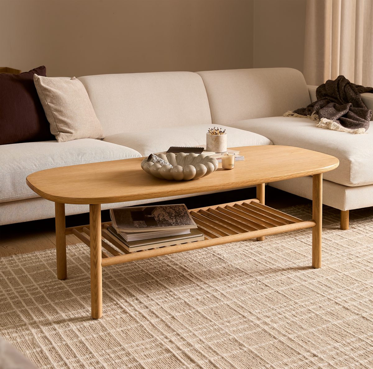 Lys stue med beige hjørnesofa og ovalt sofabord i eg med hylde; bøger og skål på bordet samt lille lys, neutrale nuancer.