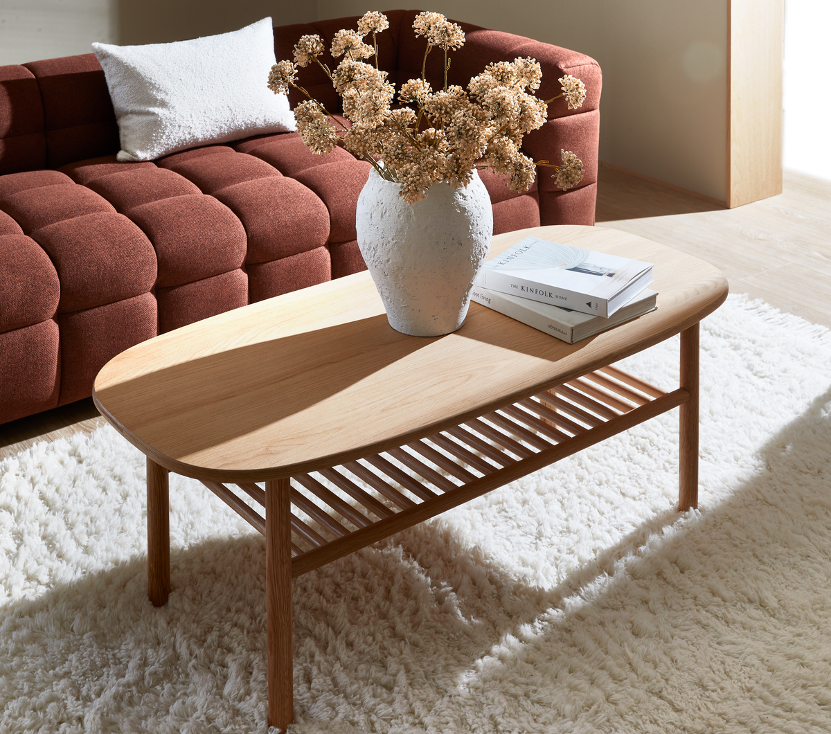 Lyst stue med ovalt sofabord i eg på en blød hvid skindagtig måtte, en hvid struktureret vase med tørrede blomster og en bogstak, der inkluderer “The Kinfolk Home”, foran en rustfarvet tufted sofa i sollys.