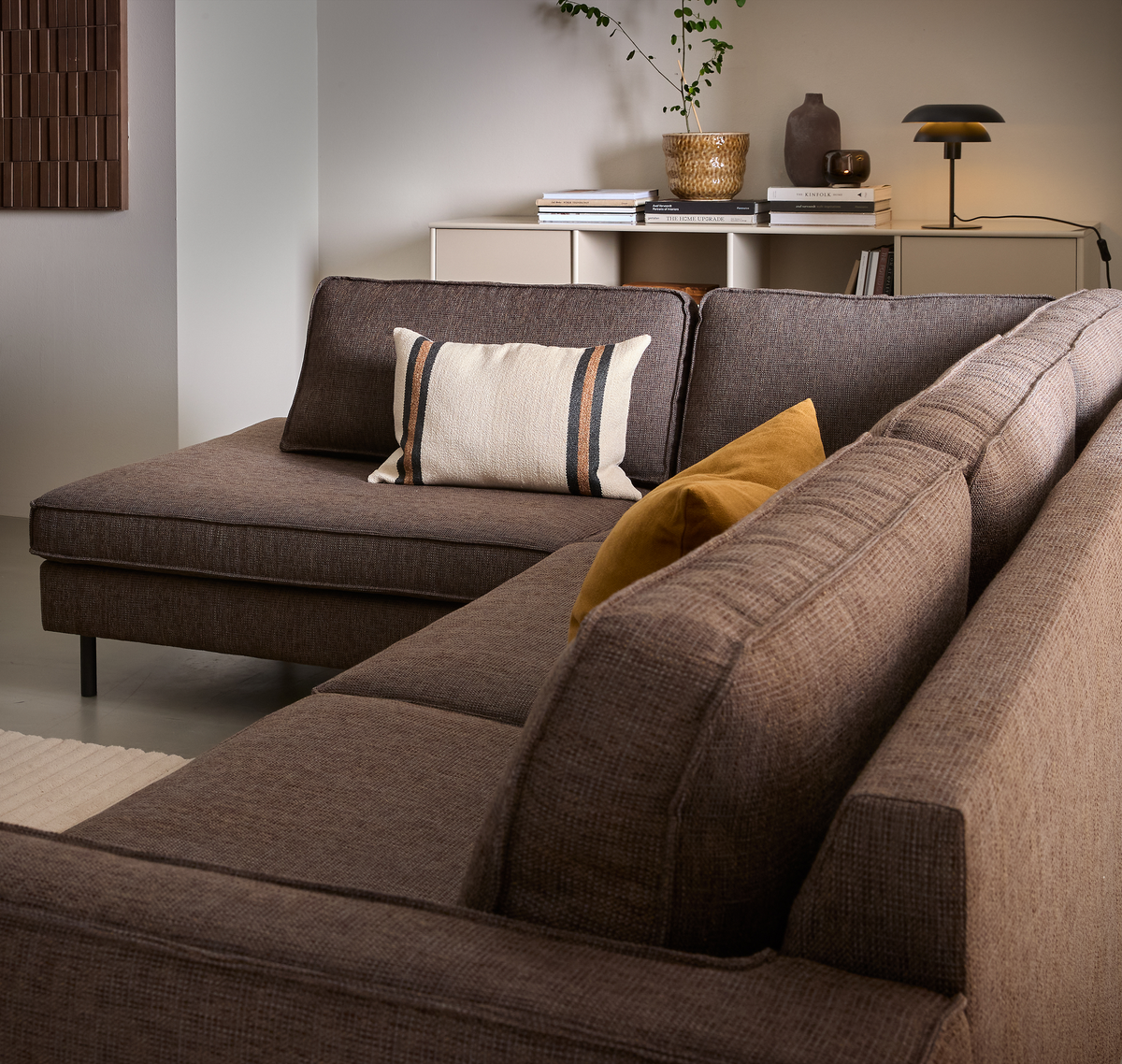 Brun stof-hjørnesofa med stribet pude og gul pude i stuen; skænk med bordlampe, bøger, plante i kurv og keramik i baggrunden.
