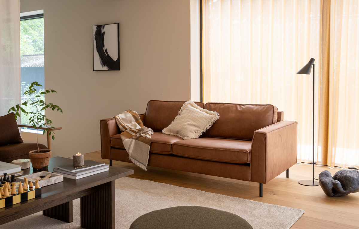 Stue med brun lædersofa, beige pude og plaid, mørkt sofabord med skakspil, bøger og tændt lys; planter, gulvlampe og lyse gardiner i dagslys.