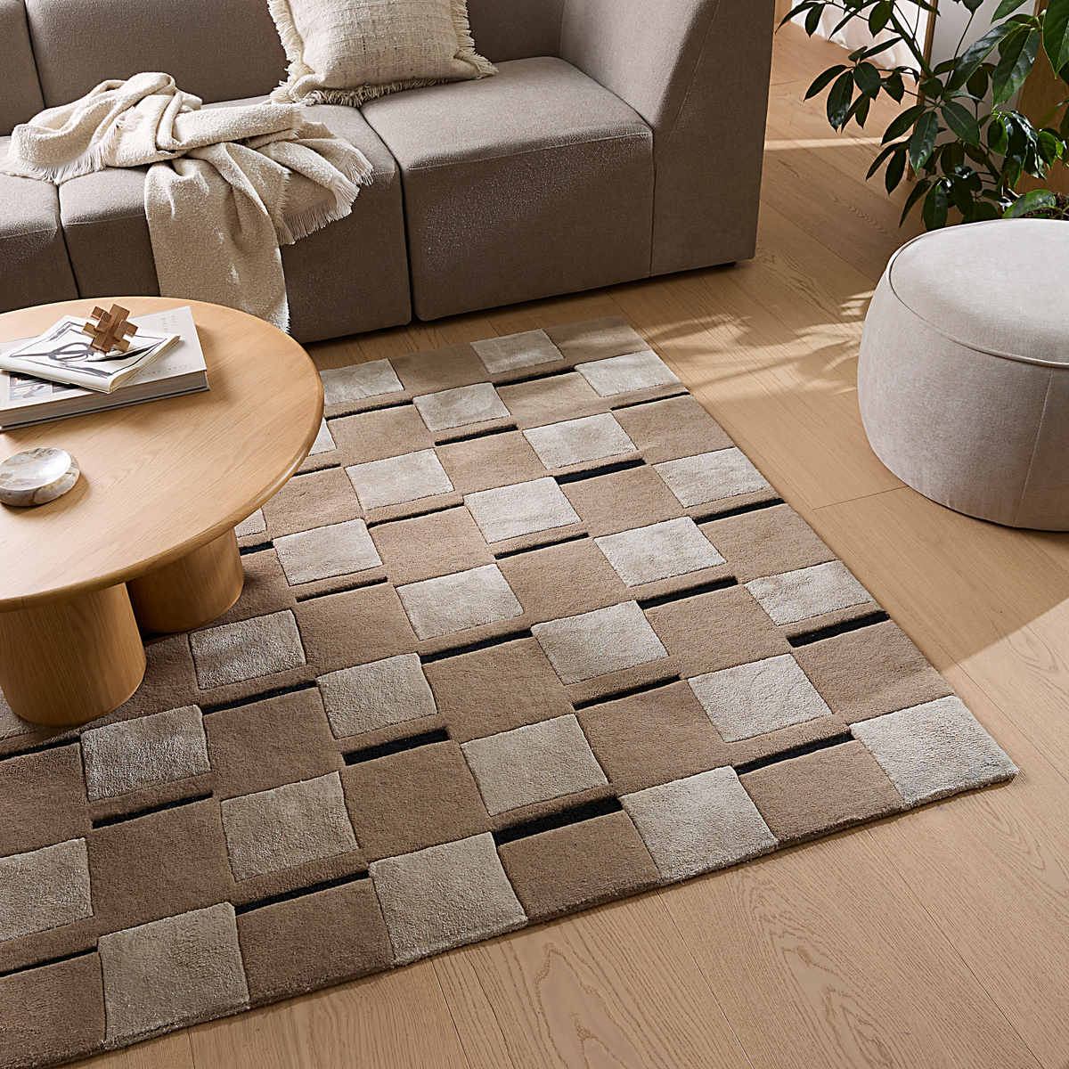 Stue i neutrale toner med ternet beige tæppe, rundt sofabord i træ med bøger og dekor, beige sofa med plaid og pude, grøn plante og puf på lyst trægulv i dagslys.