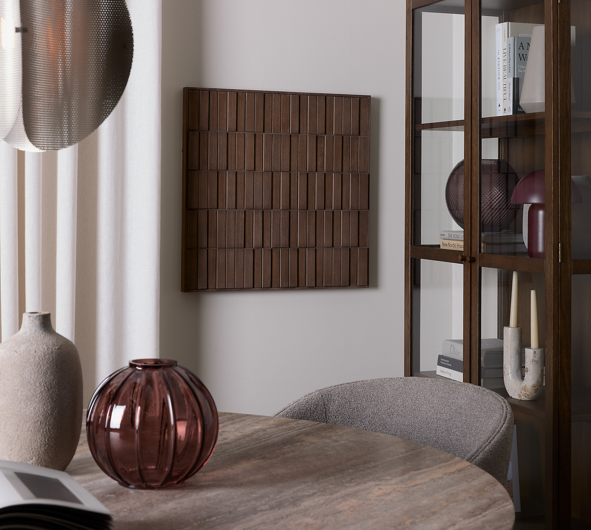 Stilrent spisestuehjørne med rundt stenbord, vinrød glasvase og beige keramikvase; grå polstret stol, vitrineskab i mørkt træ med bøger og dekorationer, træpanel som vægkunst og lyse gardiner.