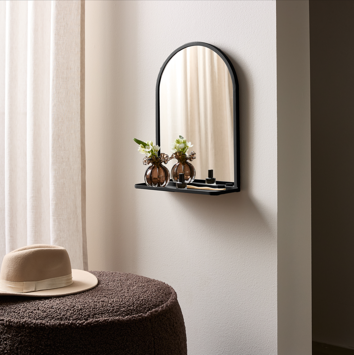 Vægspejl med sort buet ramme og lille hylde, pyntet med to brune glasvaser med blomster og en lille parfume; beige hat på en brun puf ved siden af, lyse gardiner i baggrunden.