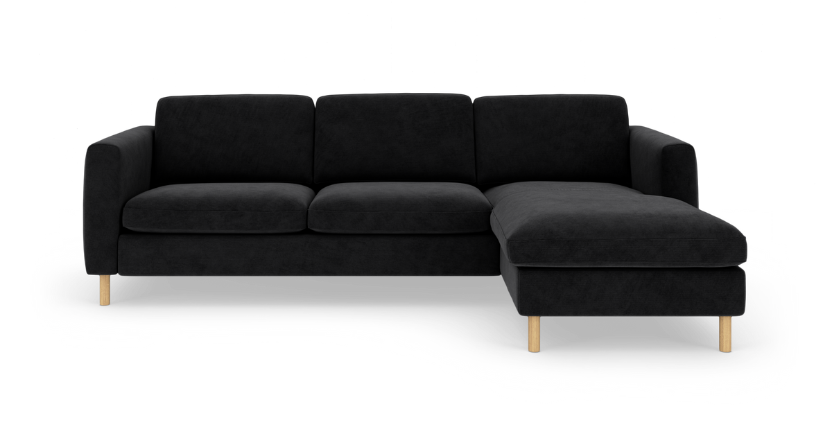 Sort hjørnesofa med chaiselong til højre og lyse træben, frilagt på hvid baggrund.