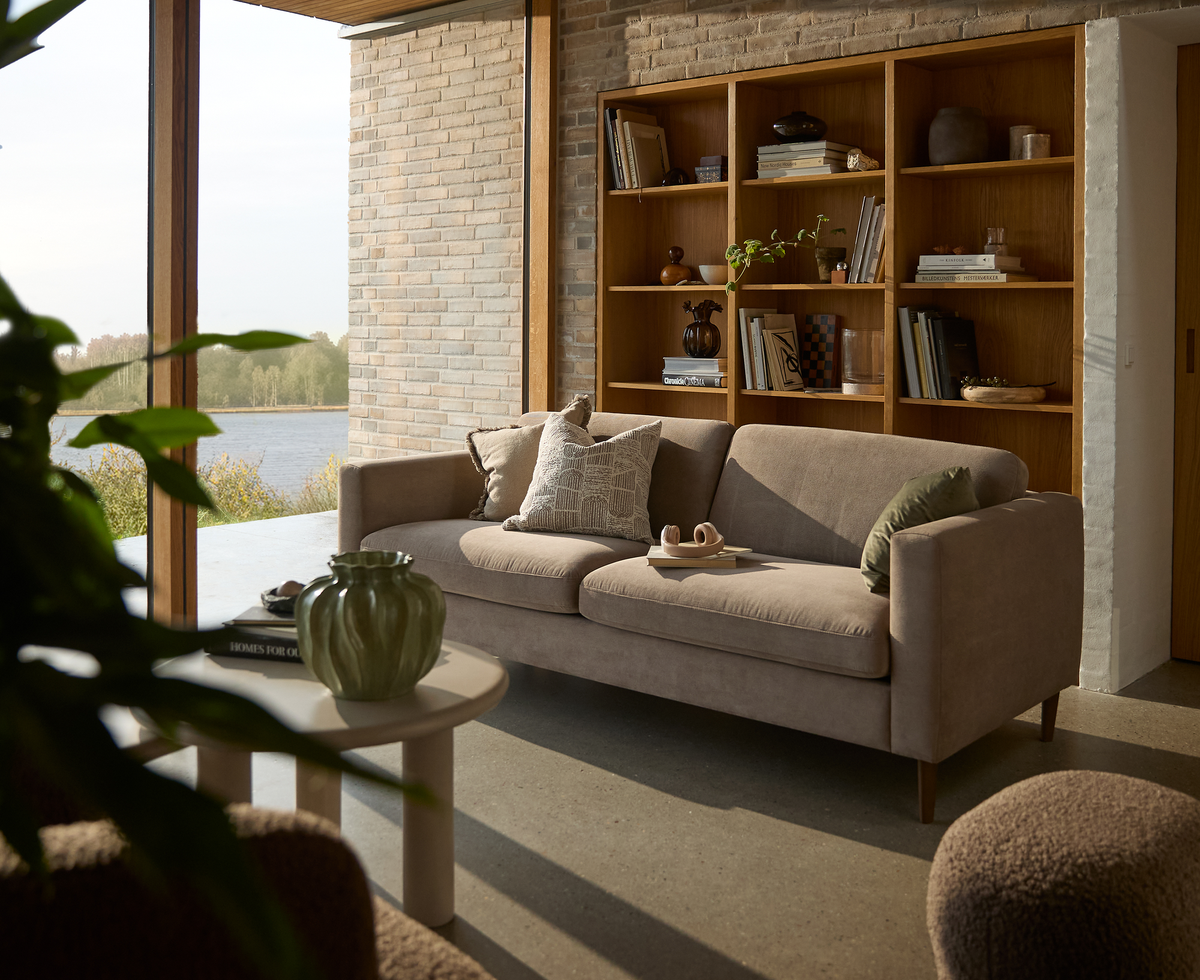 Lys skandinavisk stue med beige sofa, indbygget reol i træ, rundt sofabord med grøn vase og store vinduer med udsigt over en sø.