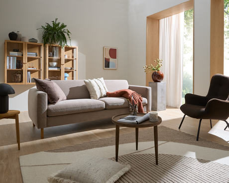 Lys skandinavisk stue med beige sofa, rundt sofabord med magasin og lille lys, neutralt tæppe og rustfarvet plaid; træhylder med bøger, plante, abstrakt kunst og stort vindue med lette gardiner i dagslys.