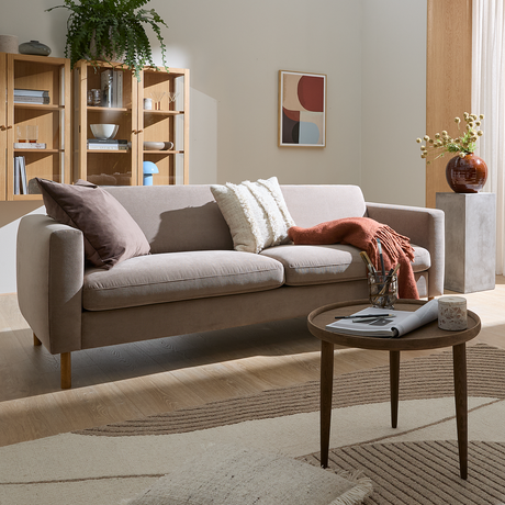 Lyst stue med beige sofa, puder og plaid, rundt sofabord med magasin og lys, stor tæppe, åbne hylder, plante og vase ved et vindue med lette gardiner.