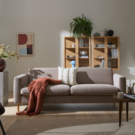 Lyst stue med beige sofa, hvid pude og rustfarvet plaid, væghængt træreol med planter og keramik, abstrakt vægkunst og blødt mønstret tæppe i dagslys.