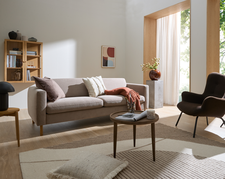 Lys stue i neutrale toner med beige sofa, brun lænestol, rundt træ-sofabord med magasin og lys, væghylde og kunst, stort vindue med lette gardiner og vase med blomster.