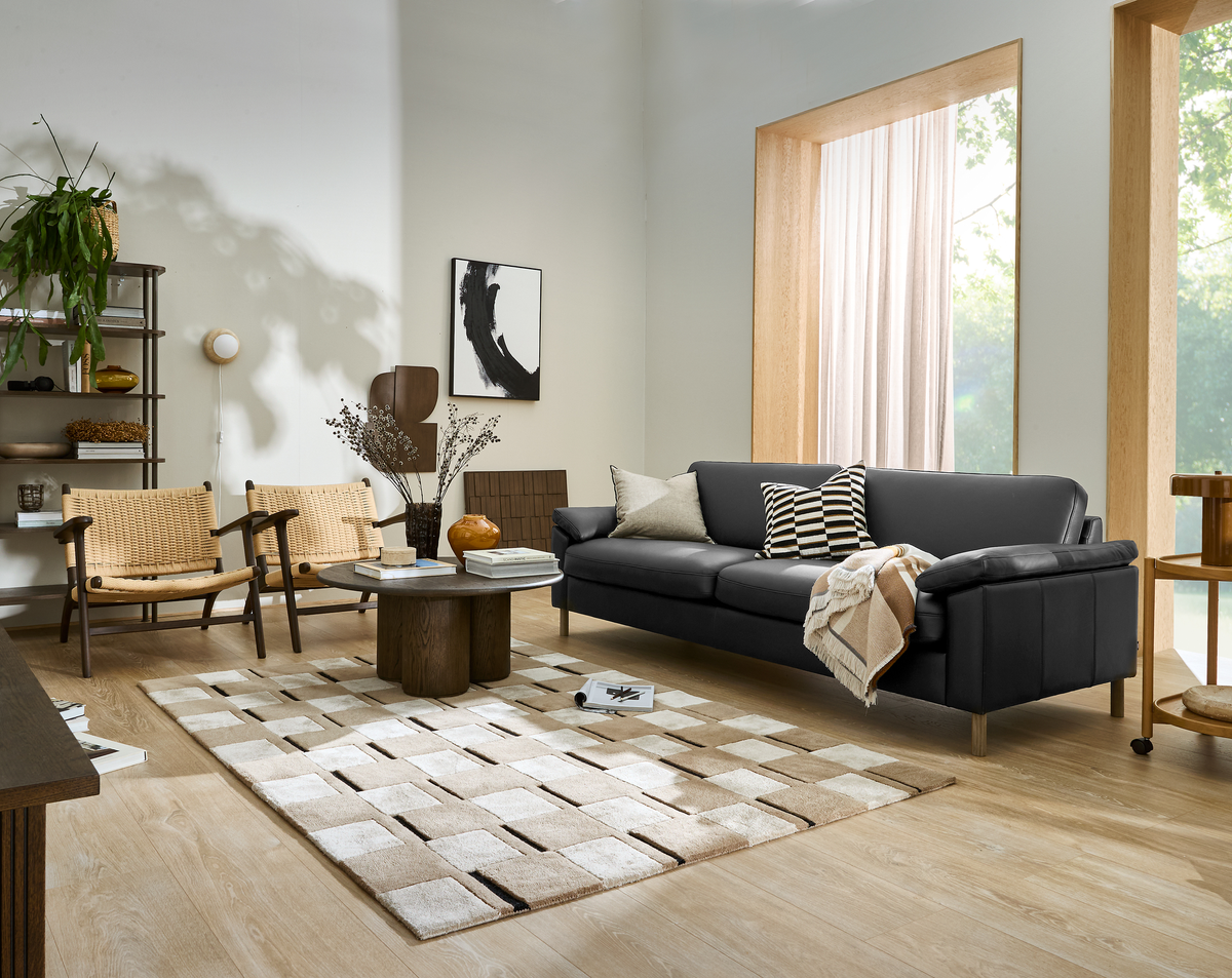 Skandinavisk stue med sort lædersofa, flettede lænestole, sofabord i træ og neutral ternet tæppe; puder og plaid på sofaen, stor vinduesniche med lette gardiner og hylde med planter i dagslys.
