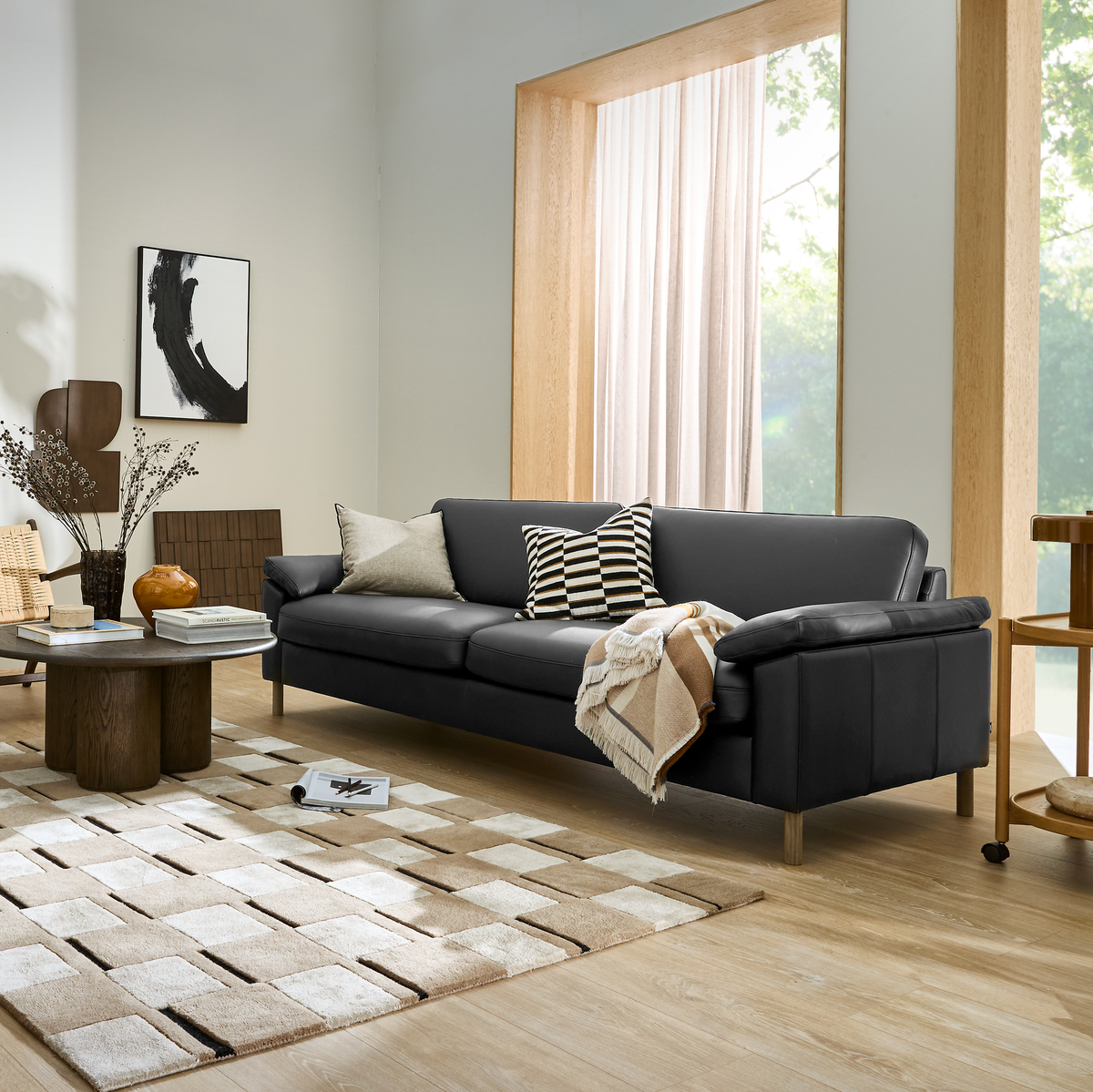 Moderne skandinavisk stue med sort sofa, geometrisk ternet tæppe, puder og plaid, træsdetaljer og stort vindue med lyse gardiner.