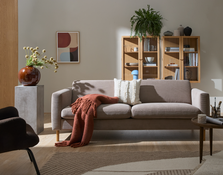 Lys stue med beige sofa, rustfarvet plaid og puder, vægmonteret træskab med bøger, dekorationer, grøn plante og lille blå bordlampe; abstrakt kunst, rundt sofabord og vase på sokkel på mønstret tæppe i dagslys.