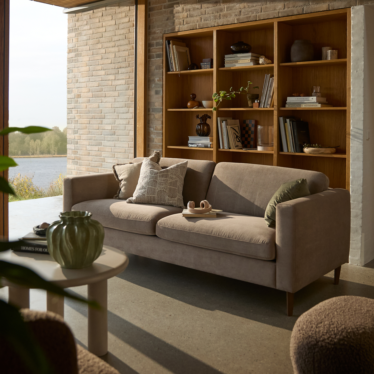 Lyst stue med beige sofa foran en træreol, sofabord med grøn vase og bogen Homes for Our Time, udsigt til en sø gennem stort vindue.