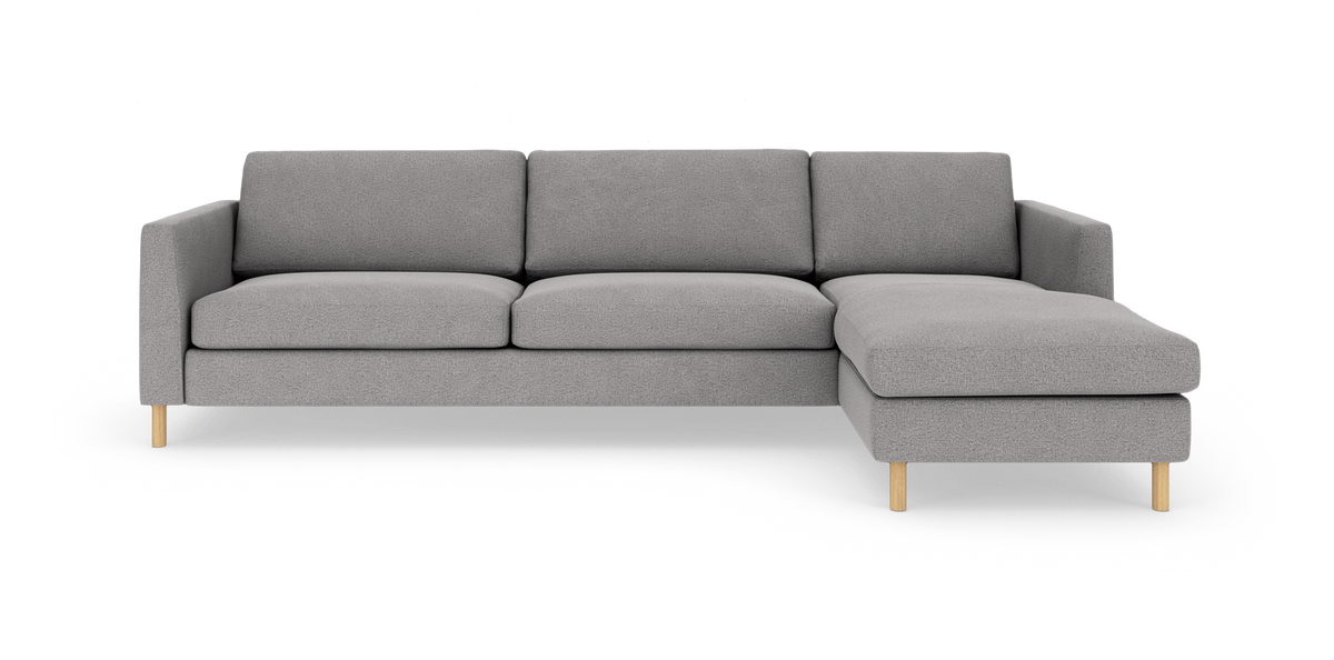 Grå hjørnesofa med chaiselong til højre, træben og bløde puder, frilagt på hvid baggrund.