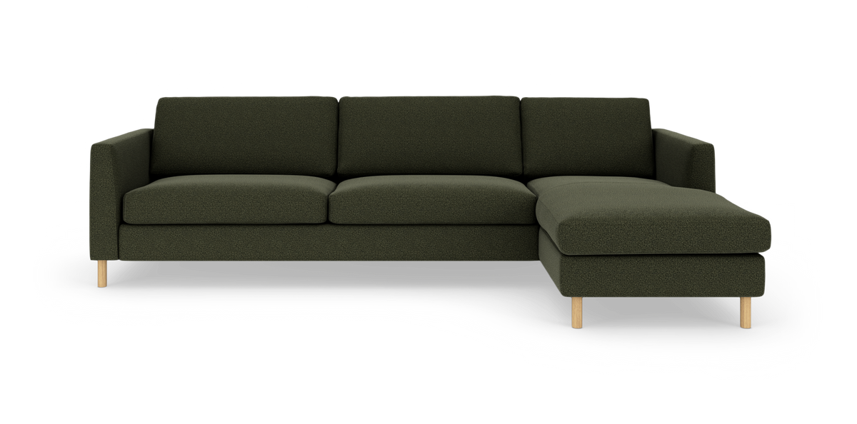 Mørkegrøn hjørnesofa med chaiselong til højre og slanke træben, set forfra mod hvid baggrund.