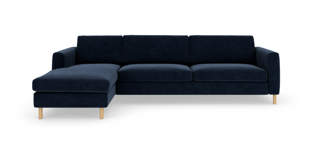 Mørkeblå hjørnesofa med venstrevendt chaiselong og lyse træben, fotograferet forfra på hvid baggrund.