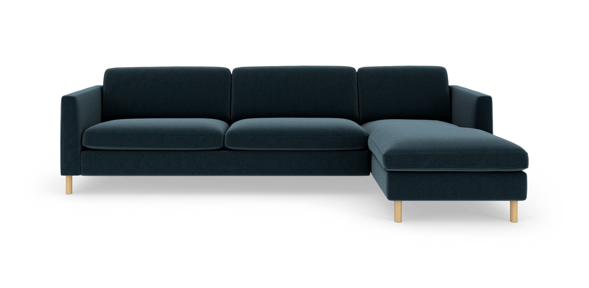 Mørkeblå sofa med chaiselong til højre og lyse træben, set forfra i studie.