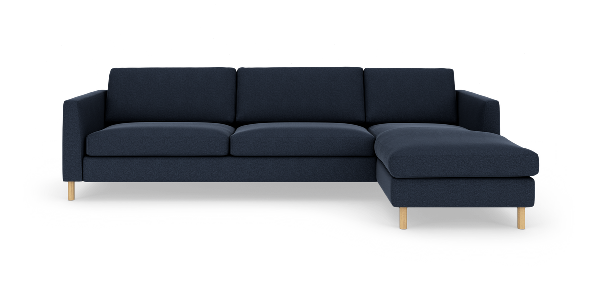 Mørkeblå hjørnesofa med chaiselong til højre og lyse træben, fritlagt på hvid baggrund.