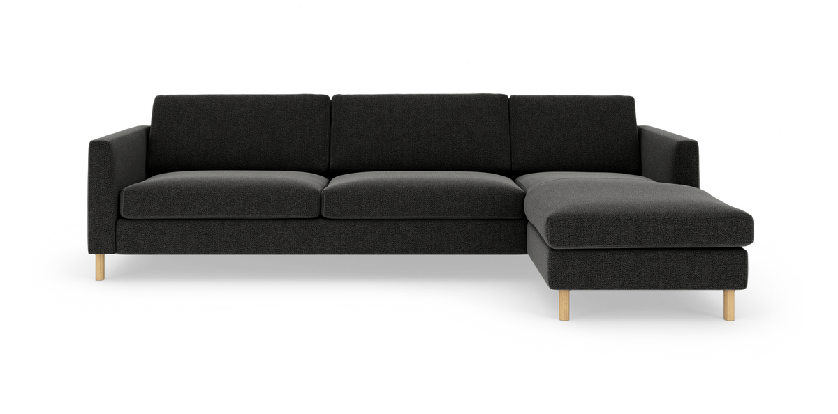 Mørkegrå hjørnesofa med chaiselong til højre og lyse træben, frilagt på hvid baggrund.