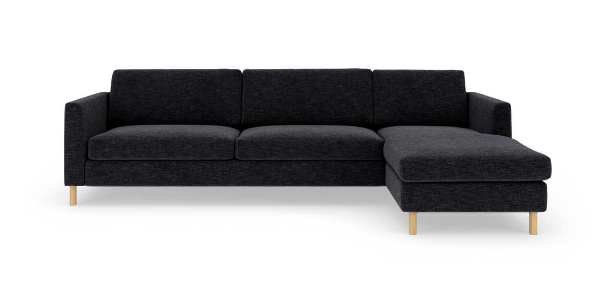 Mørkegrå hjørnesofa med chaiselong til højre og lyse træben, frilagt på hvid baggrund.