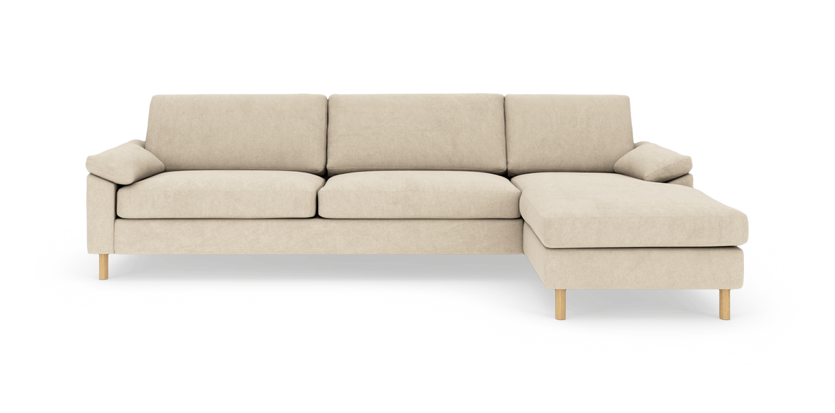 Beige hjørnesofa med chaiselong til højre, bløde puder og lyse træben, frilagt på hvid baggrund set forfra.