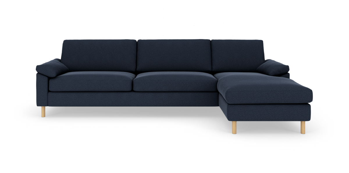 Mørkeblå hjørnesofa med chaiselong til højre og lyse træben, fotograferet forfra i studie på hvid baggrund.